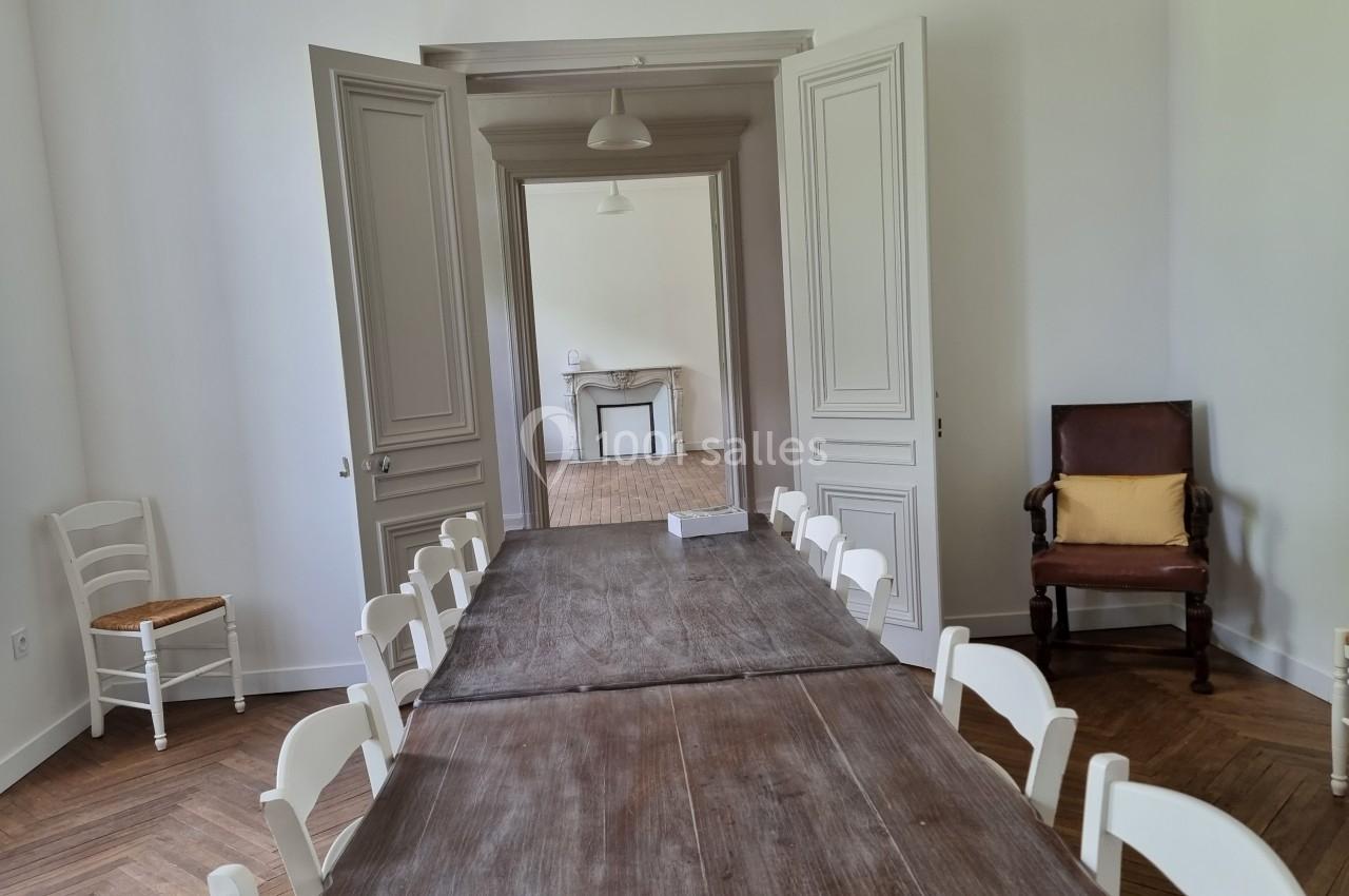 Salle lumineuse avec une grande table en bois entourée de chaises blanches, une cheminée visible en arrière-plan.