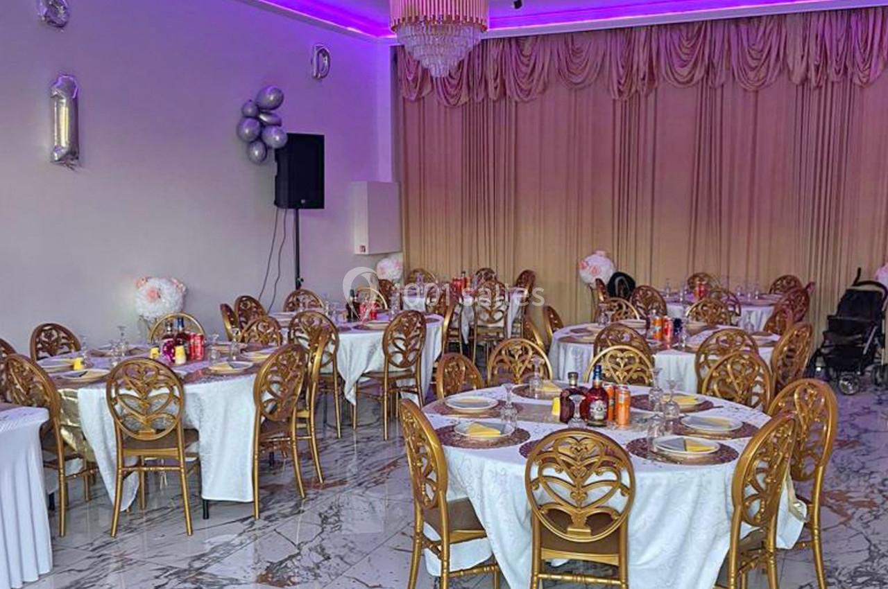 Salle de réception décorée avec des tables rondes dressées, chaises dorées et éclairage violet au plafond.