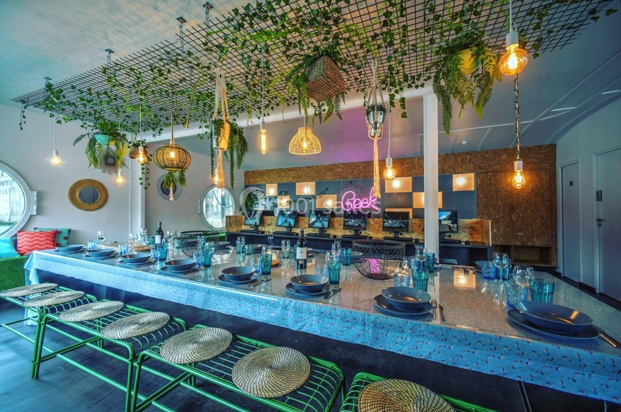 Grande table décorée avec vaisselle, entourée de chaises, sous un plafond orné de plantes suspendues et luminaires.