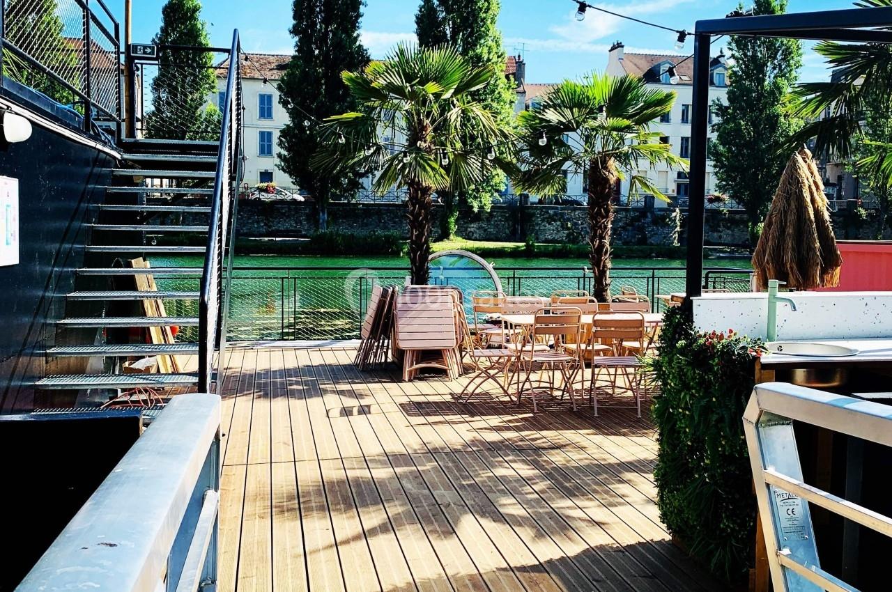 Terrasse en bois avec tables et chaises, bordée de palmiers, donnant sur un canal et des bâtiments en arrière-plan.