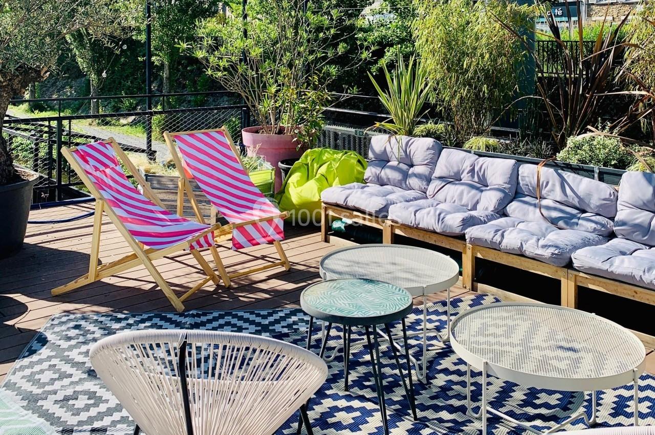 Terrasse aménagée avec chaises longues rayées, canapé à coussins, tables basses et plantes entourées de verdure.