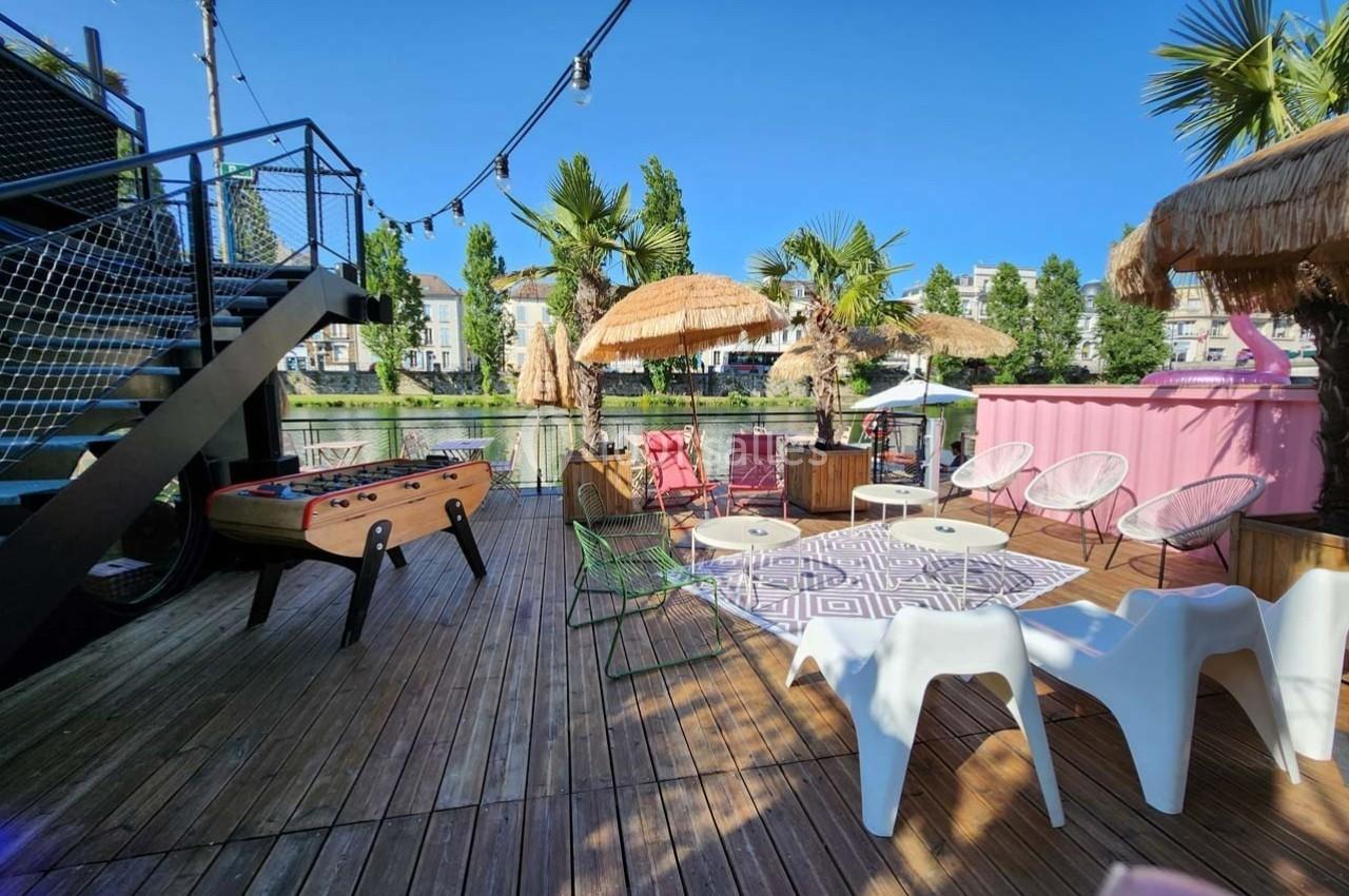 Terrasse en bois avec baby-foot, chaises, tables, parasols en paille et vue sur un canal bordé d'arbres.