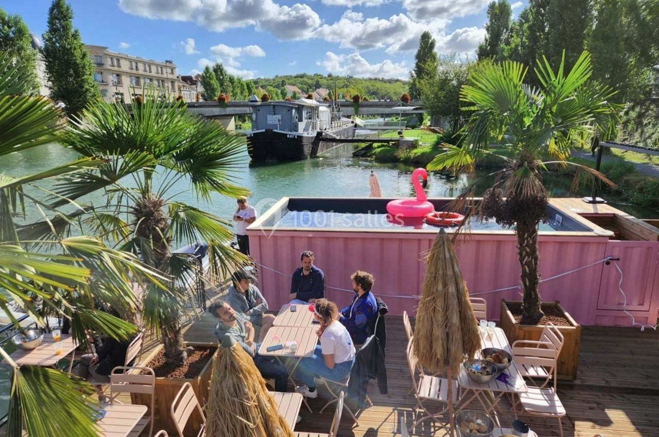 Terrasse en bois avec tables, palmiers et piscine surplombant un canal, sous un ciel partiellement nuageux.