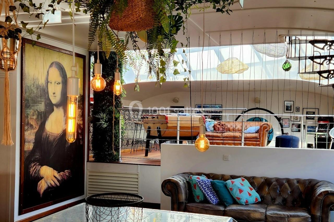 Espace intérieur lumineux avec canapé en cuir, plantes suspendues, tableau de la Joconde et baby-foot en arrière-plan.