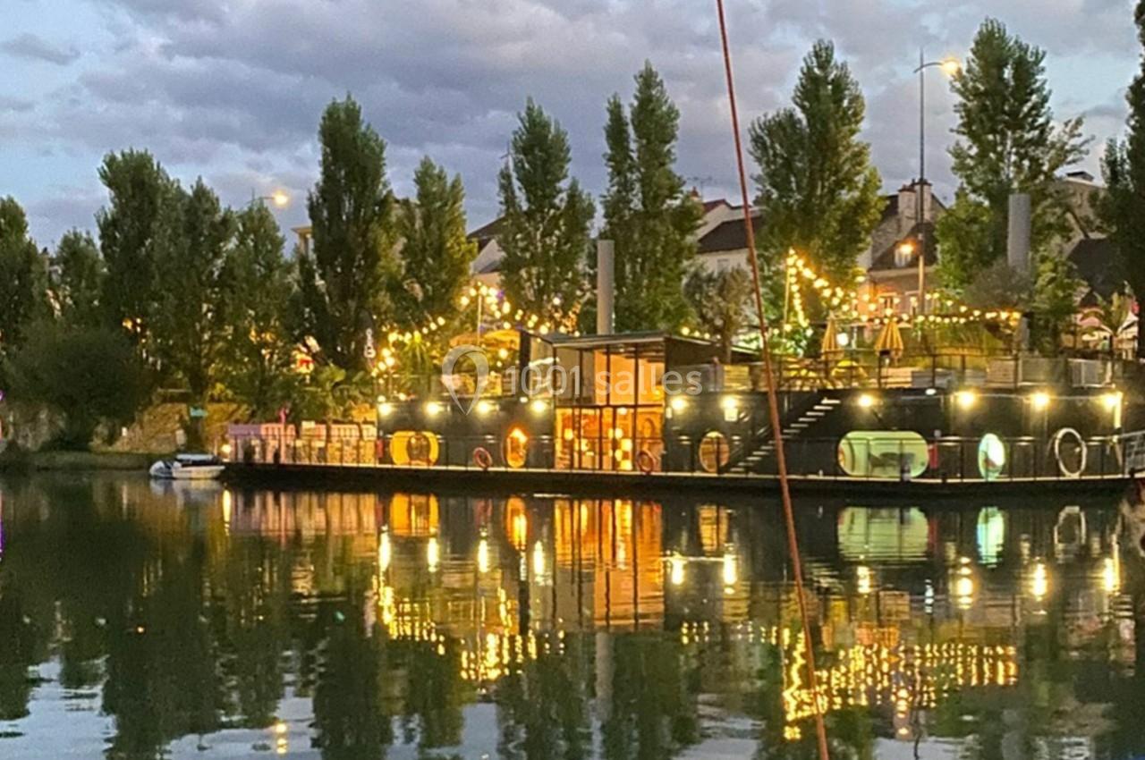Reflet d'une péniche illuminée sur un plan d'eau, entourée d'arbres et de guirlandes lumineuses au crépuscule.
