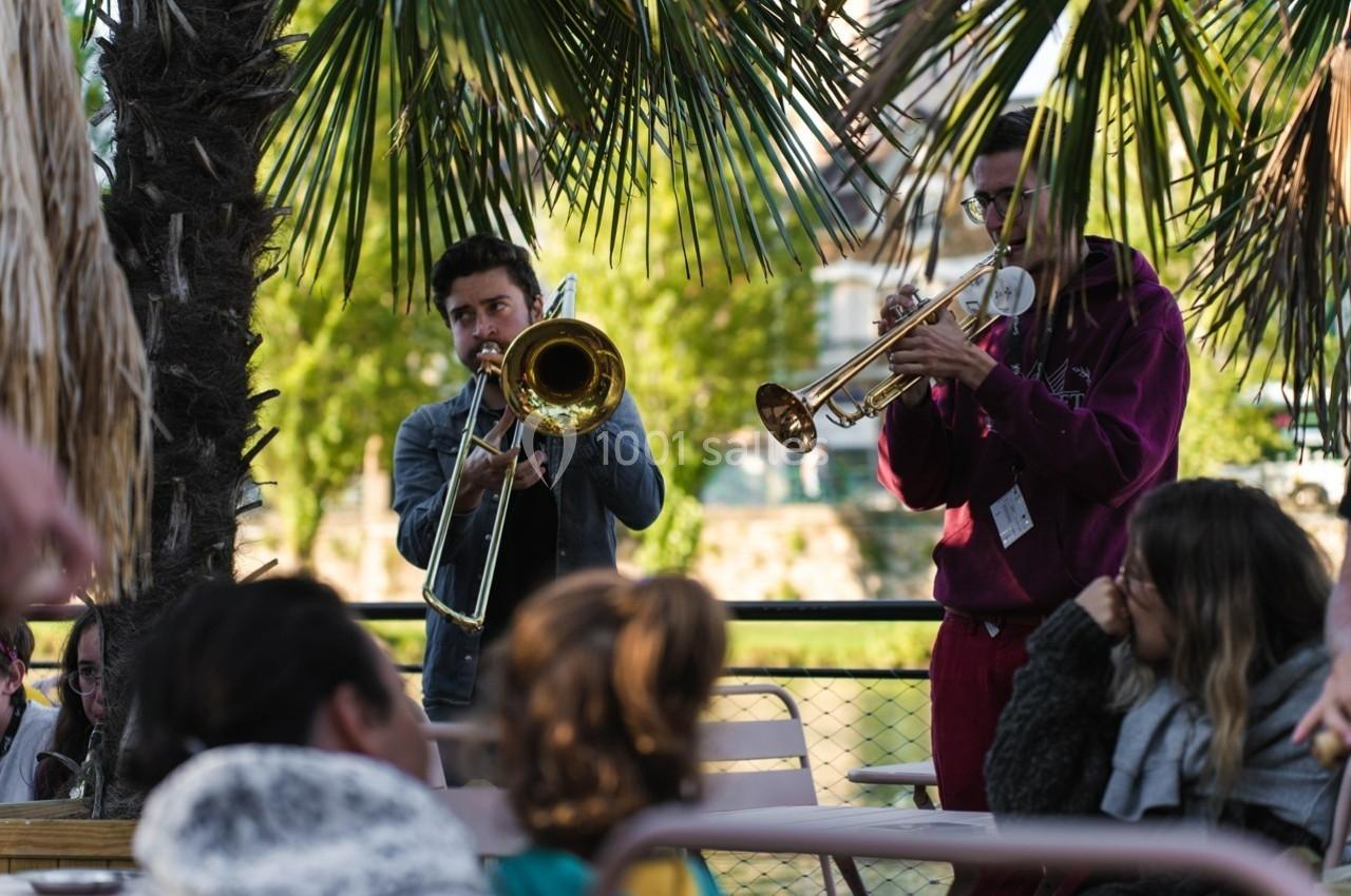 Deux musiciens jouent du trombone et de la trompette en plein air devant un petit public sous des palmiers.