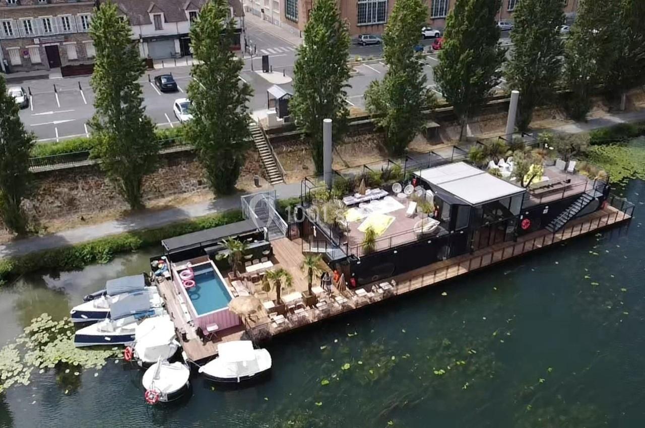 Vue aérienne d'une péniche aménagée avec terrasse, piscine et espaces de détente, amarrée sur une rivière bordée d'arbres.