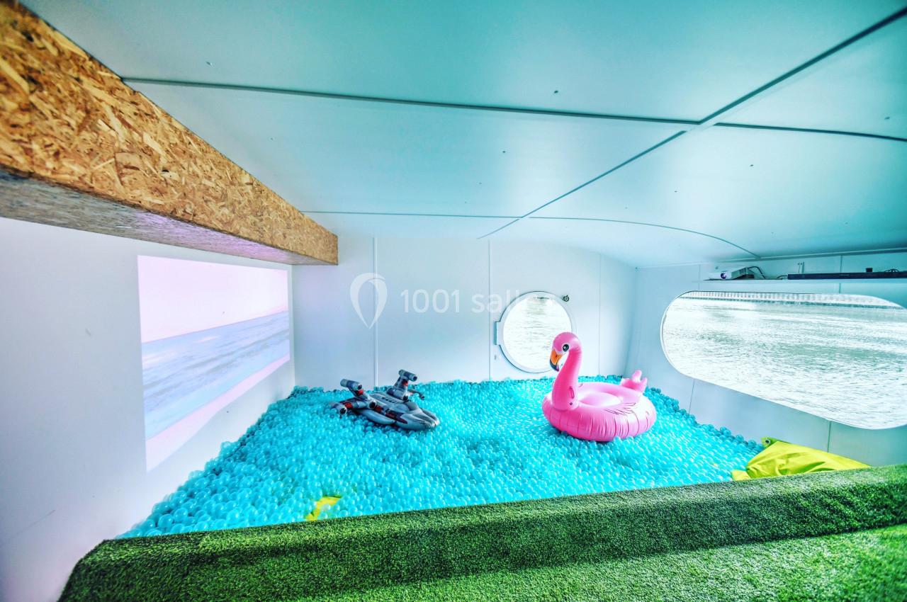 Intérieur d'une pièce avec une piscine à balles bleues, un flamant rose gonflable et une vue sur l'eau par un hublot.