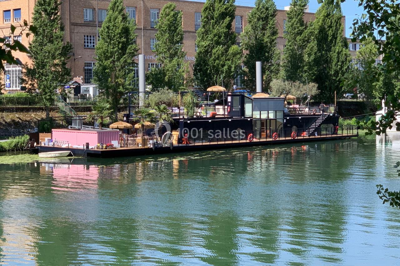 Bateau aménagé en terrasse flottante avec parasols, amarré sur une rivière bordée d'arbres et de bâtiments en arrière-plan.