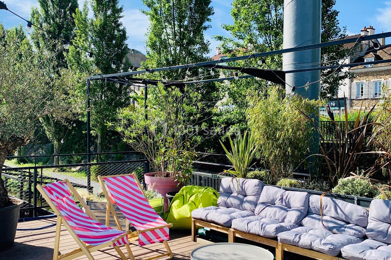 Terrasse ensoleillée avec transats colorés, canapé à coussins, tables basses et végétation environnante.
