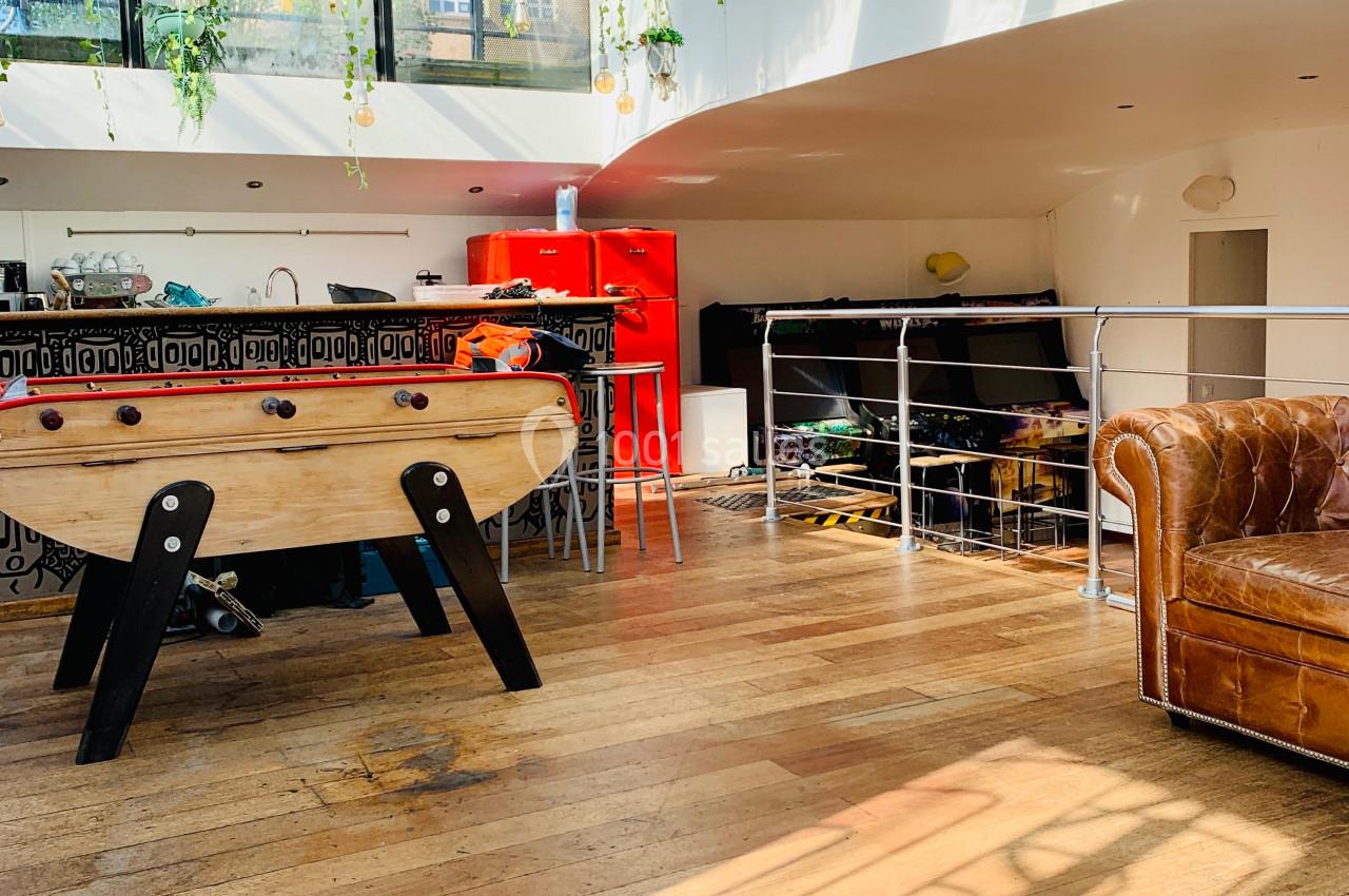 Espace lumineux avec parquet, baby-foot, canapé en cuir, cuisine rouge et verrière laissant entrer la lumière naturelle.
