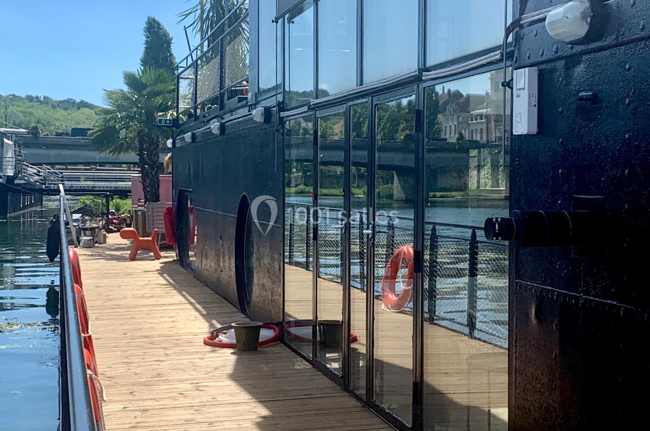 Vue latérale d'une péniche avec terrasse en bois, baies vitrées et bouées de sauvetage, amarrée au bord d'une rivière.
