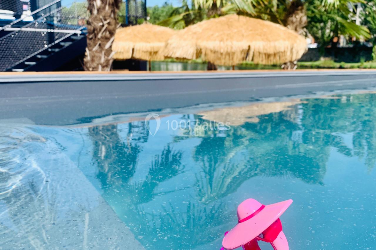 Un flamant rose miniature flotte dans une piscine entourée de palmiers et d'une cabane au toit de chaume.