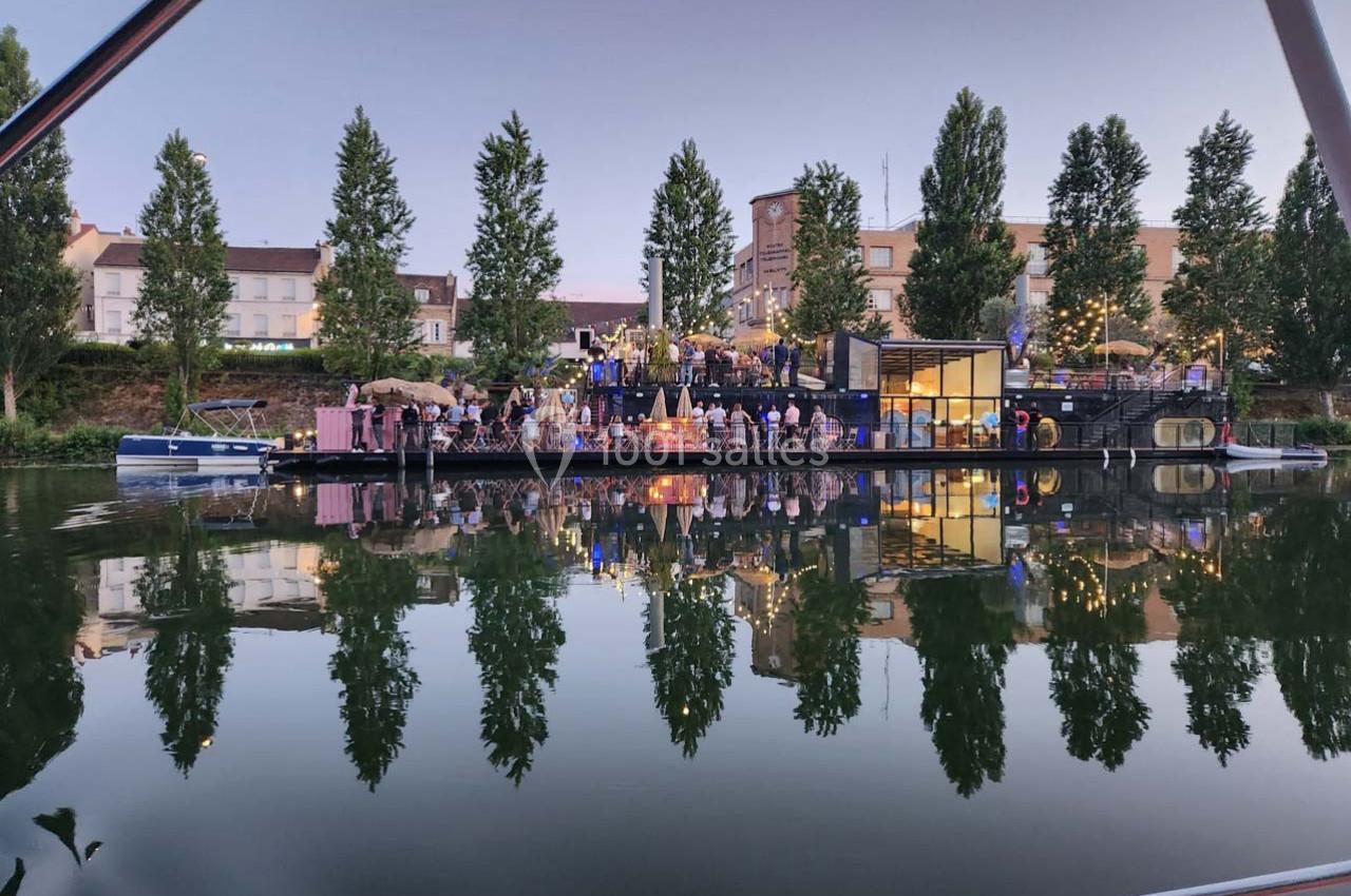 Personnes rassemblées sur une péniche éclairée au crépuscule, amarrée près d'arbres et de bâtiments au bord de l'eau.