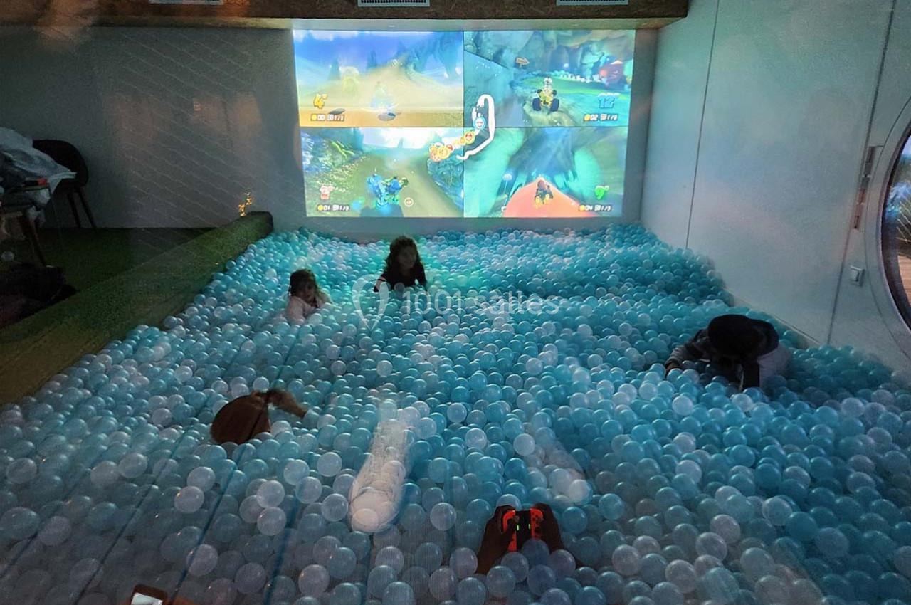 Des enfants jouent dans une piscine à balles bleues devant un écran projetant un jeu vidéo multijoueur.