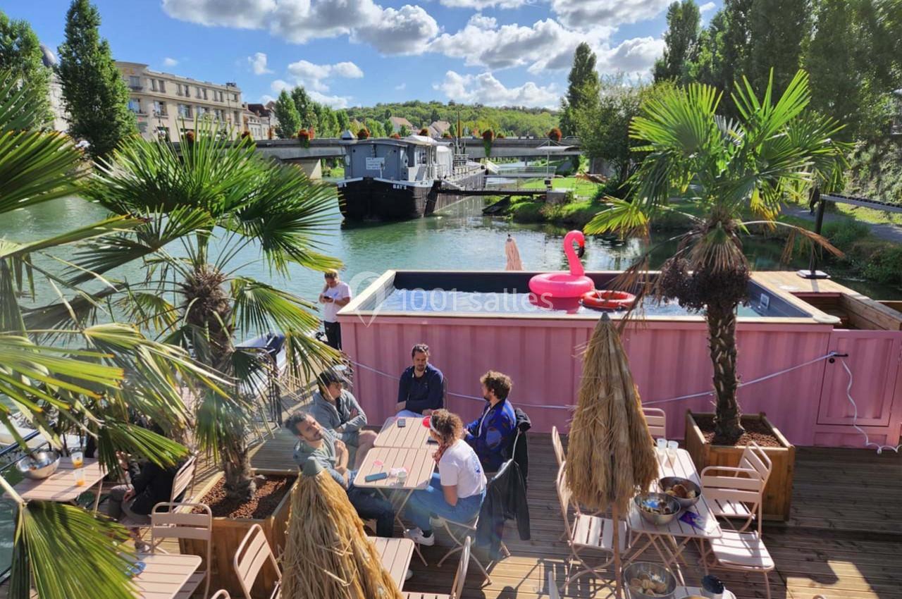 Terrasse ensoleillée avec tables, palmiers, piscine rose et vue sur une rivière bordée de végétation.