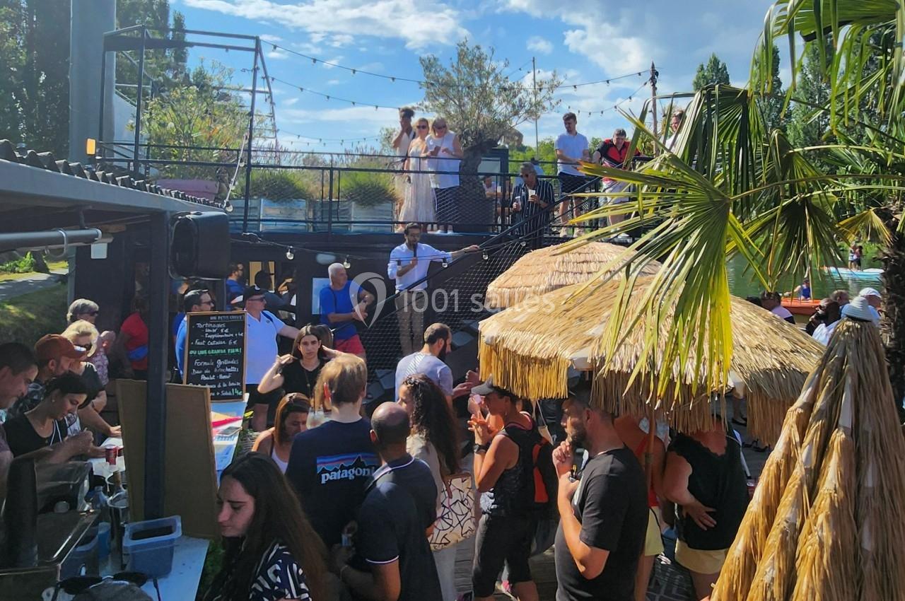 Groupe de personnes réunies dans un bar extérieur animé avec terrasse, palmiers et guirlandes lumineuses.