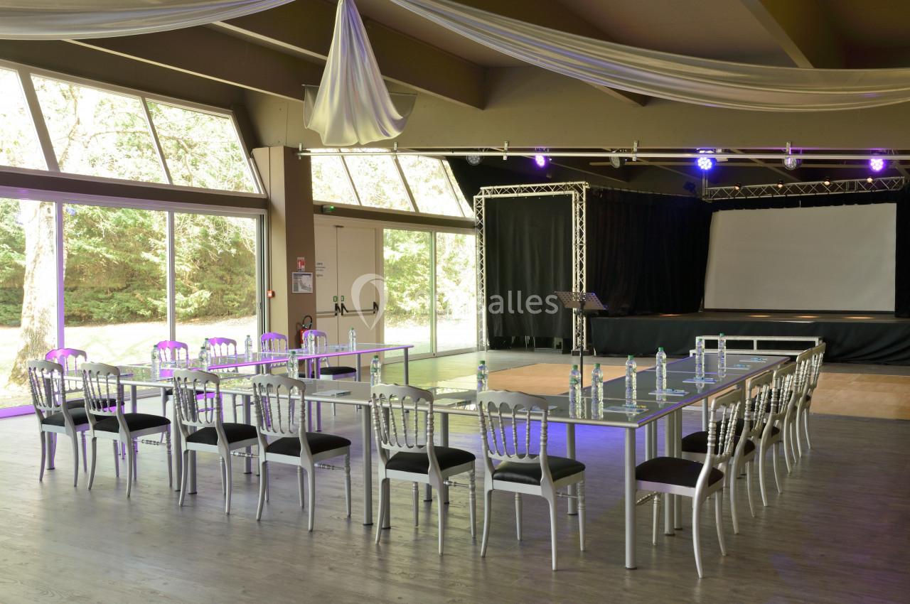 Salle de conférence lumineuse avec tables en U, chaises argentées, projecteur et scène équipée d’un écran.