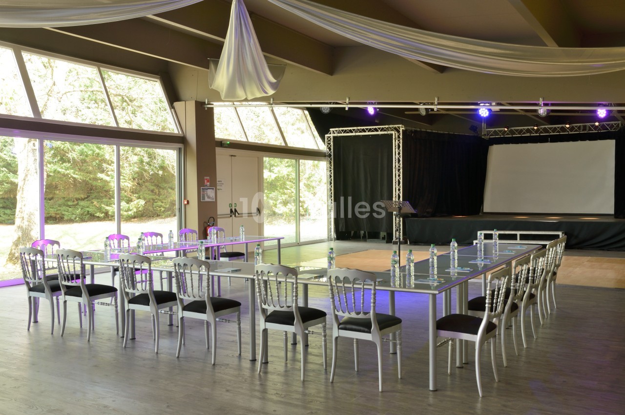 Salle de conférence lumineuse avec tables en U, chaises argentées, projecteur et scène équipée d’un écran.