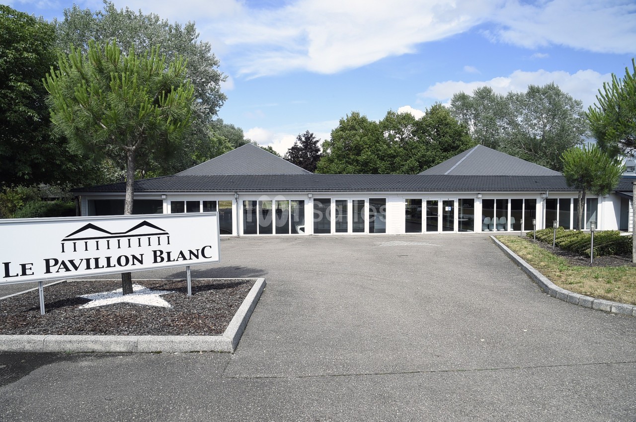 Bâtiment moderne avec toit gris, grandes baies vitrées et panneau indiquant ’Le Pavillon Blanc’ à l'entrée.