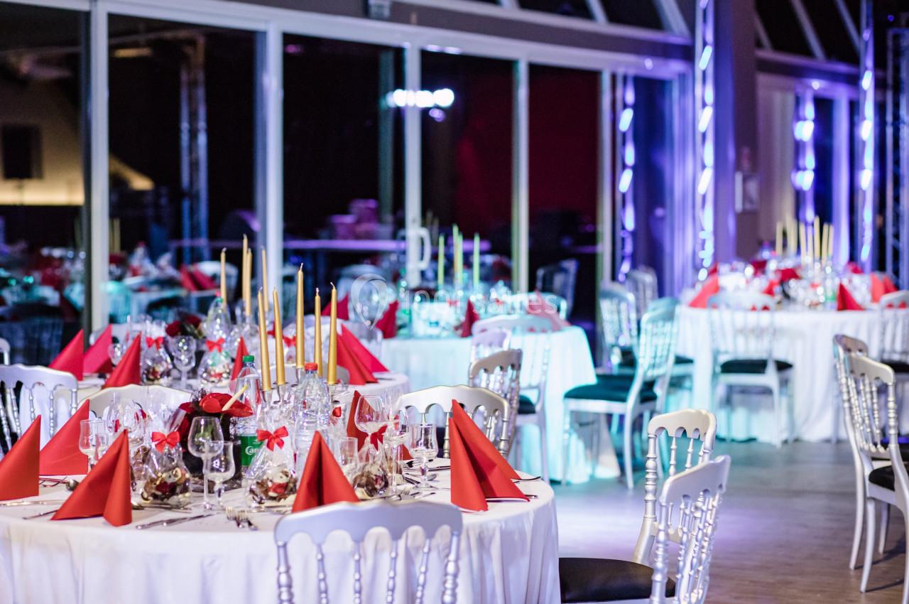 Salle de réception décorée avec des tables rondes, nappes blanches, serviettes rouges et chandeliers dorés.