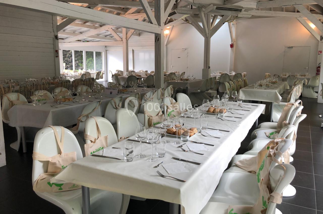 Salle de réception lumineuse avec des tables dressées, nappes blanches, chaises modernes et sacs en tissu suspendus.