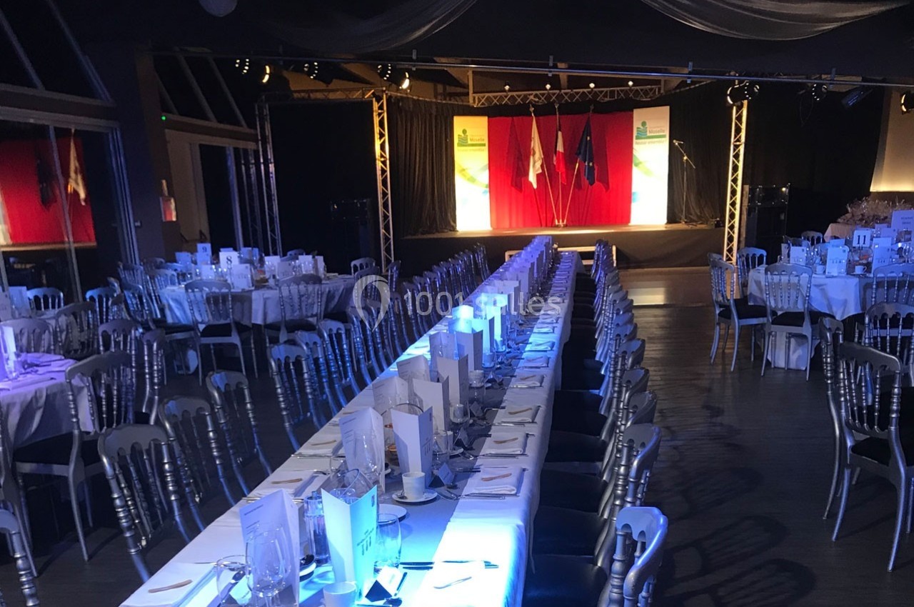 Salle de réception avec tables dressées, chaises argentées et une scène équipée de rideaux et éclairages.