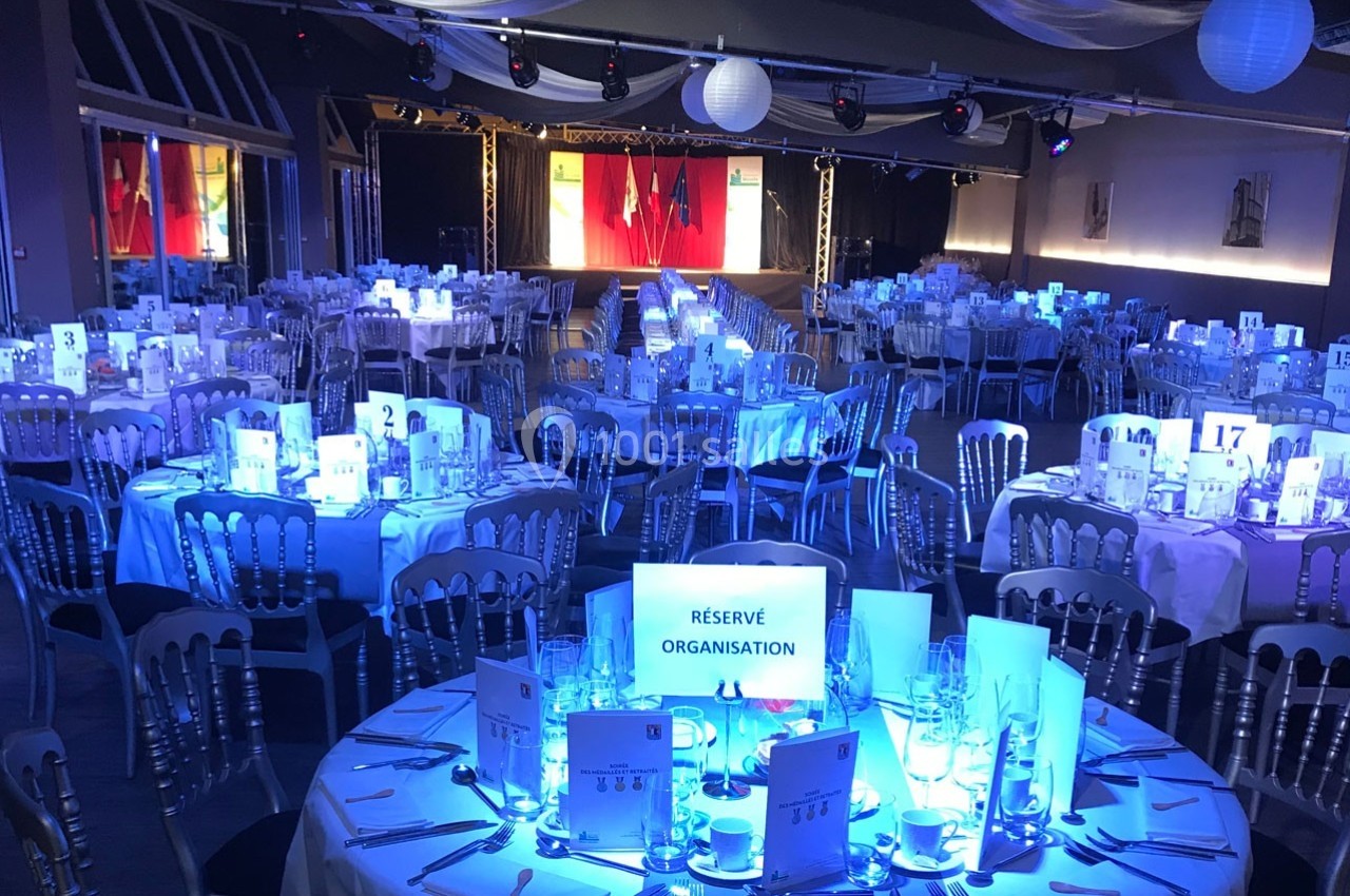 Salle de réception illuminée en bleu, avec des tables rondes dressées et un panneau ’Réservé Organisation’ visible.