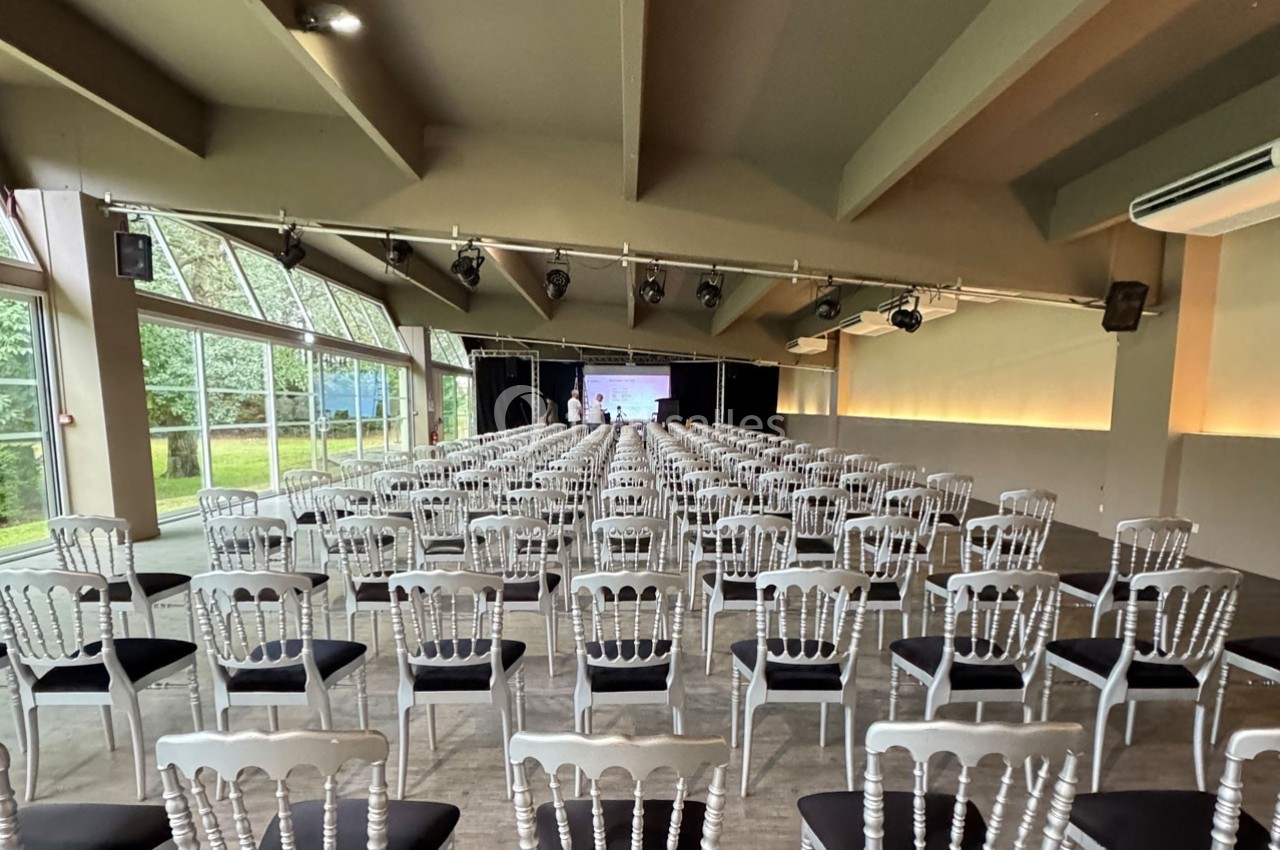 Salle de conférence lumineuse avec des rangées de chaises blanches alignées face à un écran de projection.