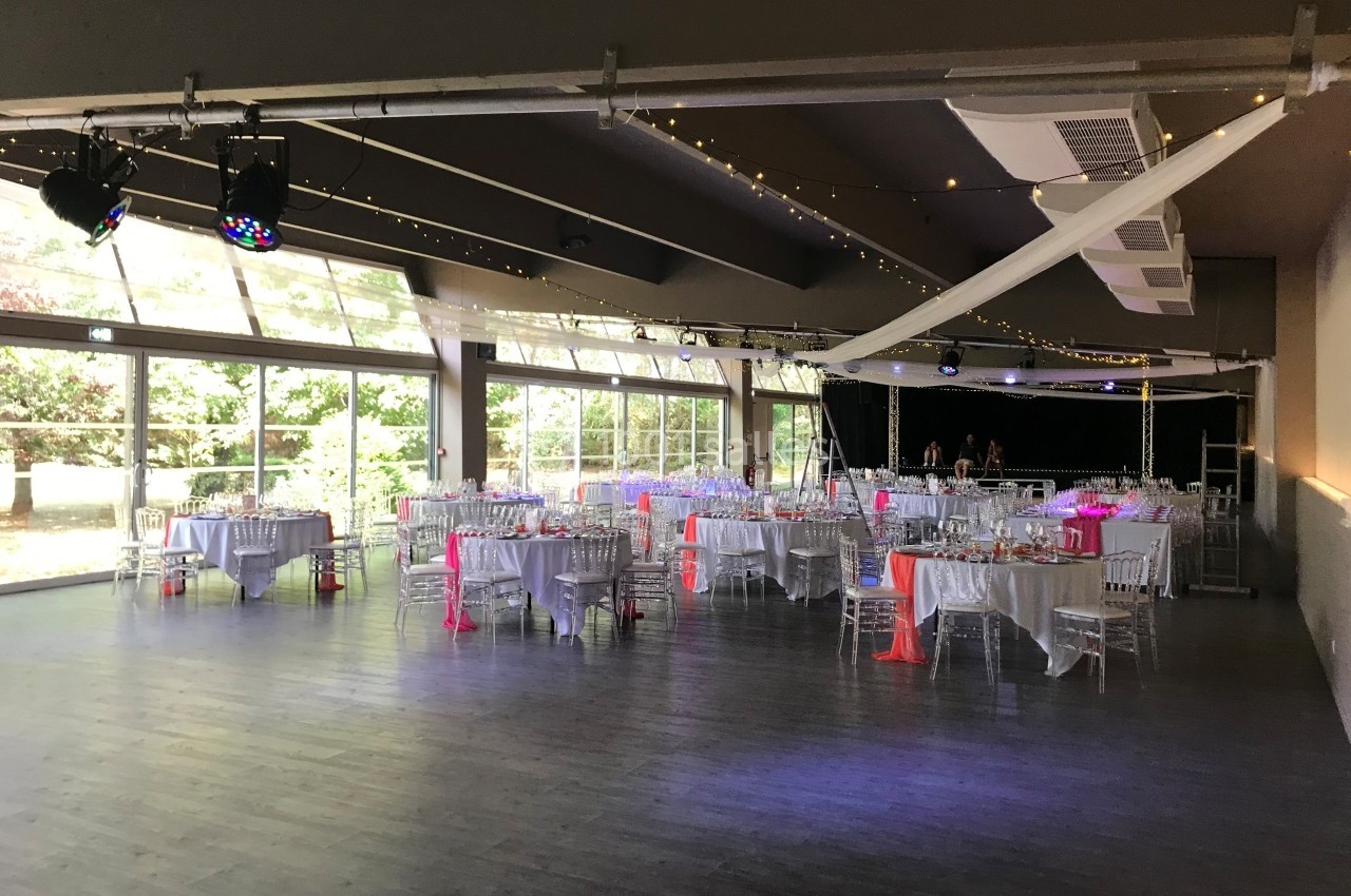 Salle de réception lumineuse avec grandes baies vitrées, tables dressées et décorations roses et blanches.