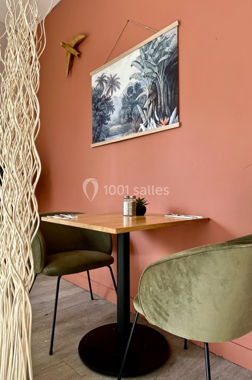 Table en bois avec deux chaises vertes, décorée d'un tableau tropical et séparée par un paravent en branches.
