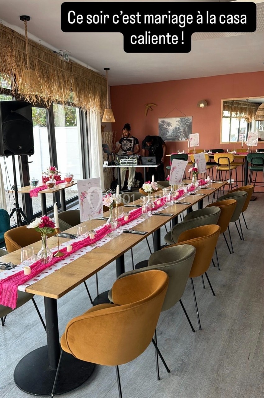 Salle décorée pour un mariage avec tables alignées, nappes roses, fleurs et chaises colorées dans un cadre chaleureux.