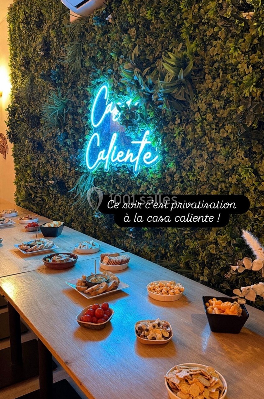 Table garnie de plats apéritifs variés devant un mur végétal avec un néon ’Casa Caliente’.