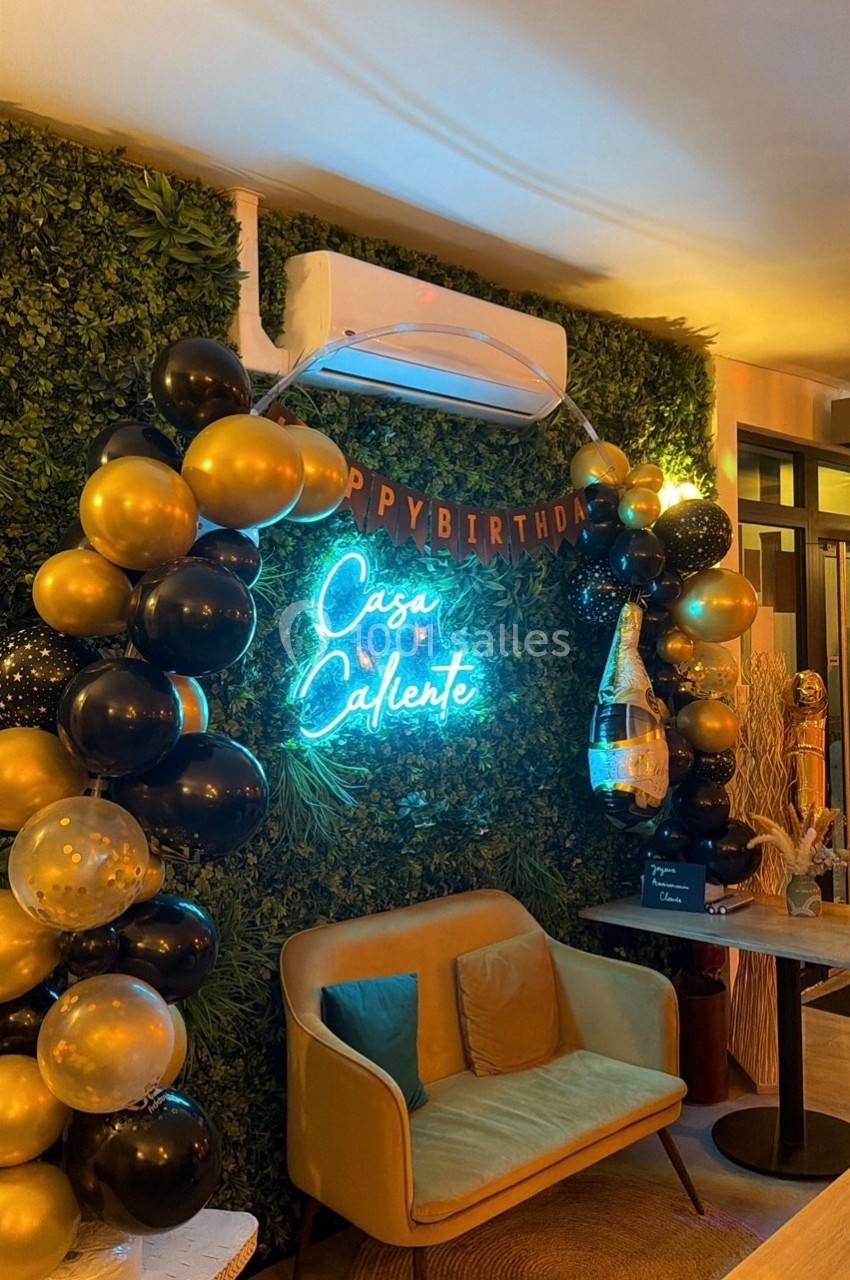 Mur végétal décoré avec des ballons noirs et dorés, néon ’Casa Caliente’, fauteuil et table dans un espace intérieur.