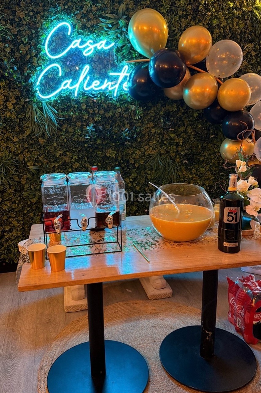 Table avec boissons, punch dans un grand bol, décor de ballons noir et or, mur végétal et néon ’Casa Caliente’.