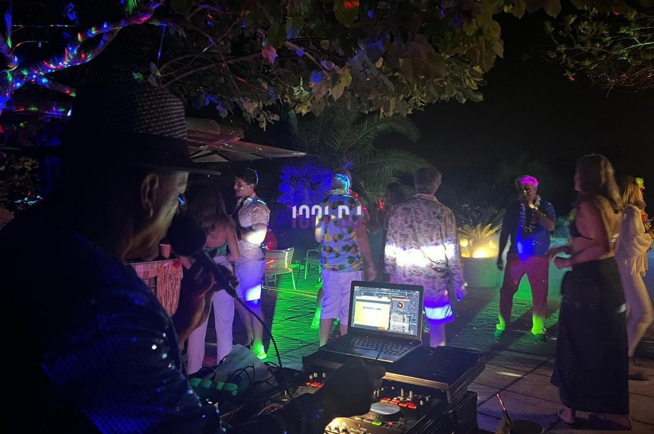 Un DJ anime une soirée en plein air avec des lumières colorées et des invités dansant sous les arbres.
