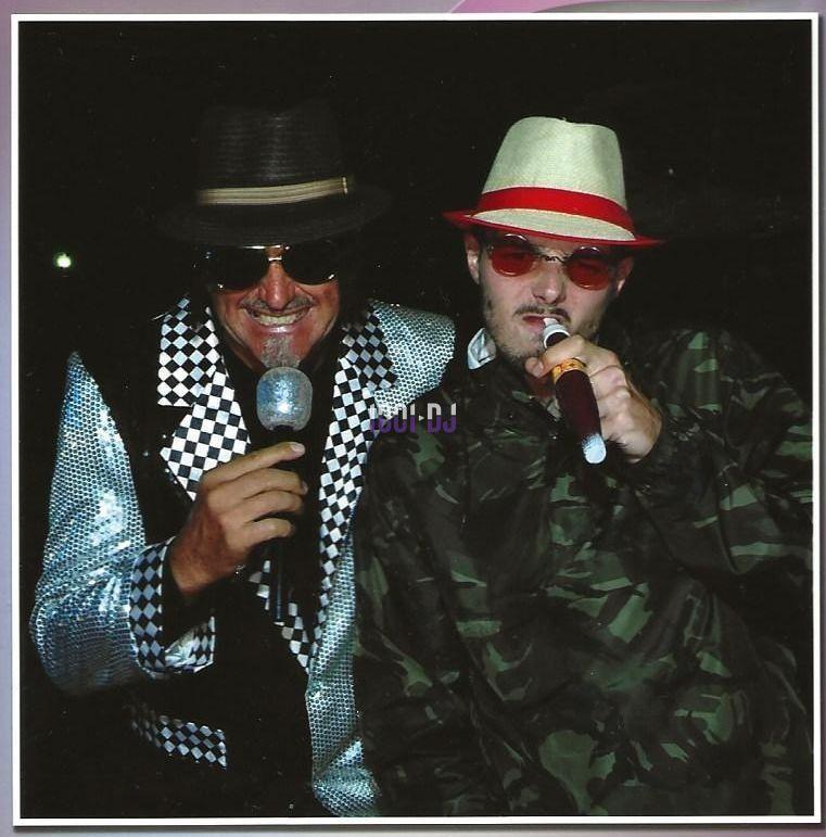 Deux hommes portant des chapeaux et des lunettes, tenant des micros, dans un environnement sombre.