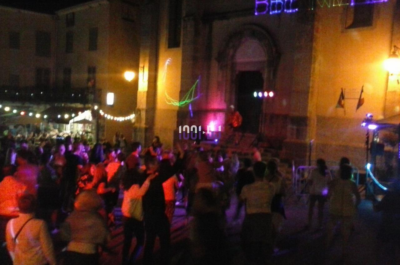 Foule dansant de nuit sur une place éclairée, devant une église avec des lumières colorées.