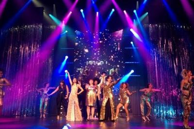 Des artistes en costumes brillants dansent sur une scène illuminée par des projecteurs colorés et un écran lumineux.