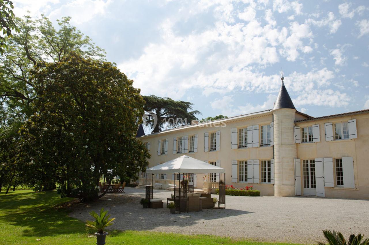 Façade d'un château en pierre avec tours, entouré d'arbres, terrasse aménagée avec parasol au premier plan.