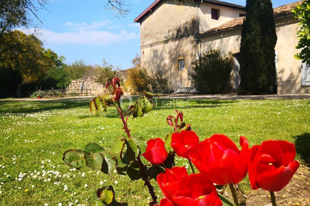 Roses rouges en premier plan dans un jardin verdoyant, avec une maison en pierre et un ciel bleu en arrière-plan.