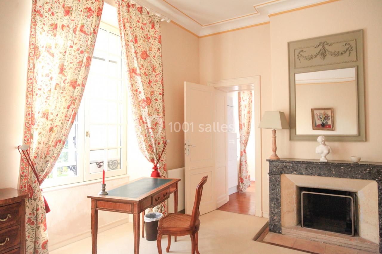 Pièce lumineuse avec bureau en bois, cheminée, miroir, rideaux fleuris et une porte ouverte sur une autre pièce.
