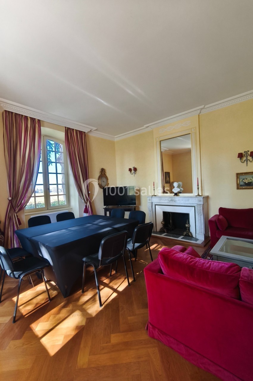 Salle de réunion lumineuse avec table noire, chaises, cheminée, canapé rouge et grandes fenêtres avec rideaux.