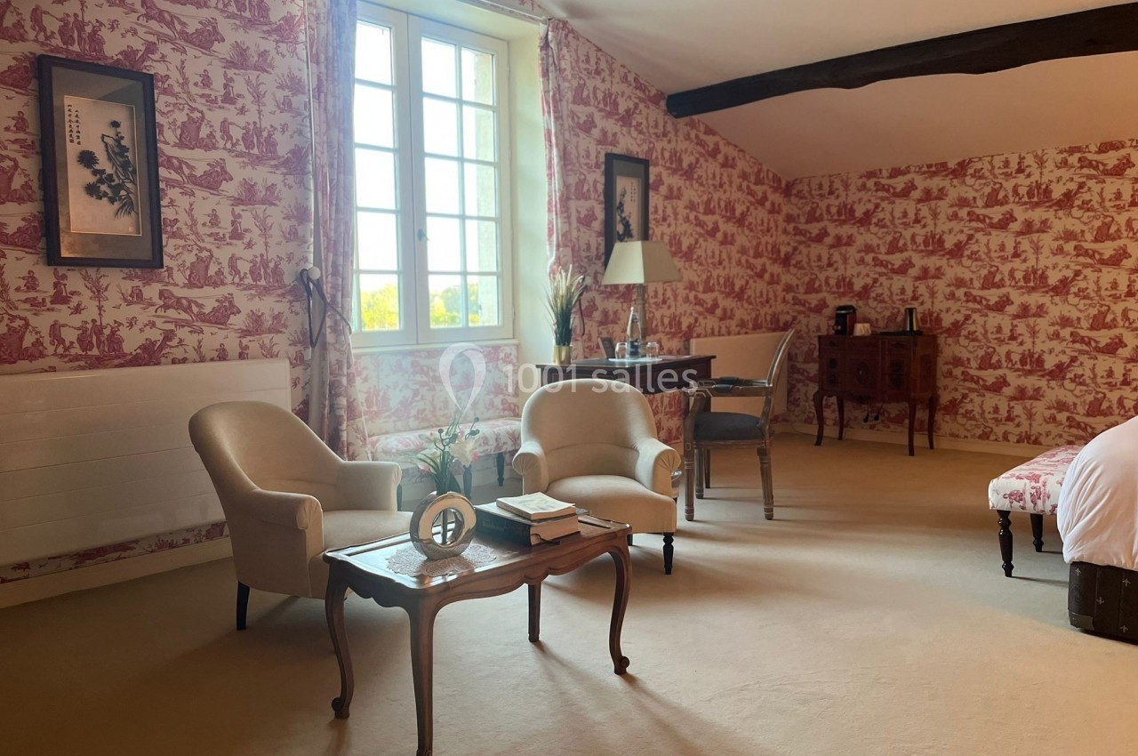 Chambre lumineuse avec murs en toile de Jouy rouge, mobilier classique, table basse et fauteuils près d'une fenêtre.