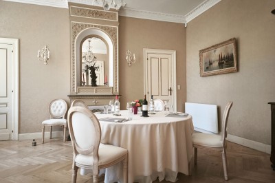 Façade claire avec volets blancs, lanterne en fer forgé et jardinières de fleurs roses en premier plan.