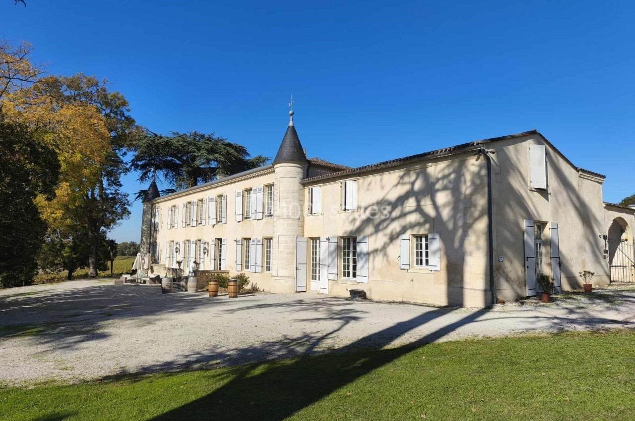 Grande maison en pierre avec tour centrale, volets blancs et jardin, éclairée par un ciel bleu dégagé.