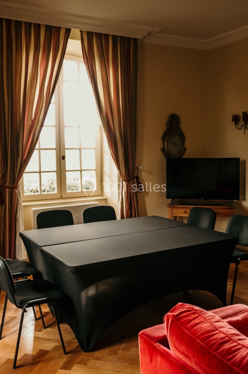 Salle avec une table noire recouverte, six chaises, un téléviseur, des rideaux rayés et une fenêtre lumineuse.