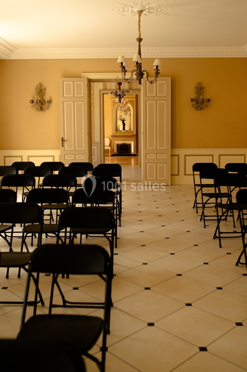 Salle lumineuse avec des chaises noires pliantes alignées, portes ouvertes sur une pièce avec cheminée et miroir.