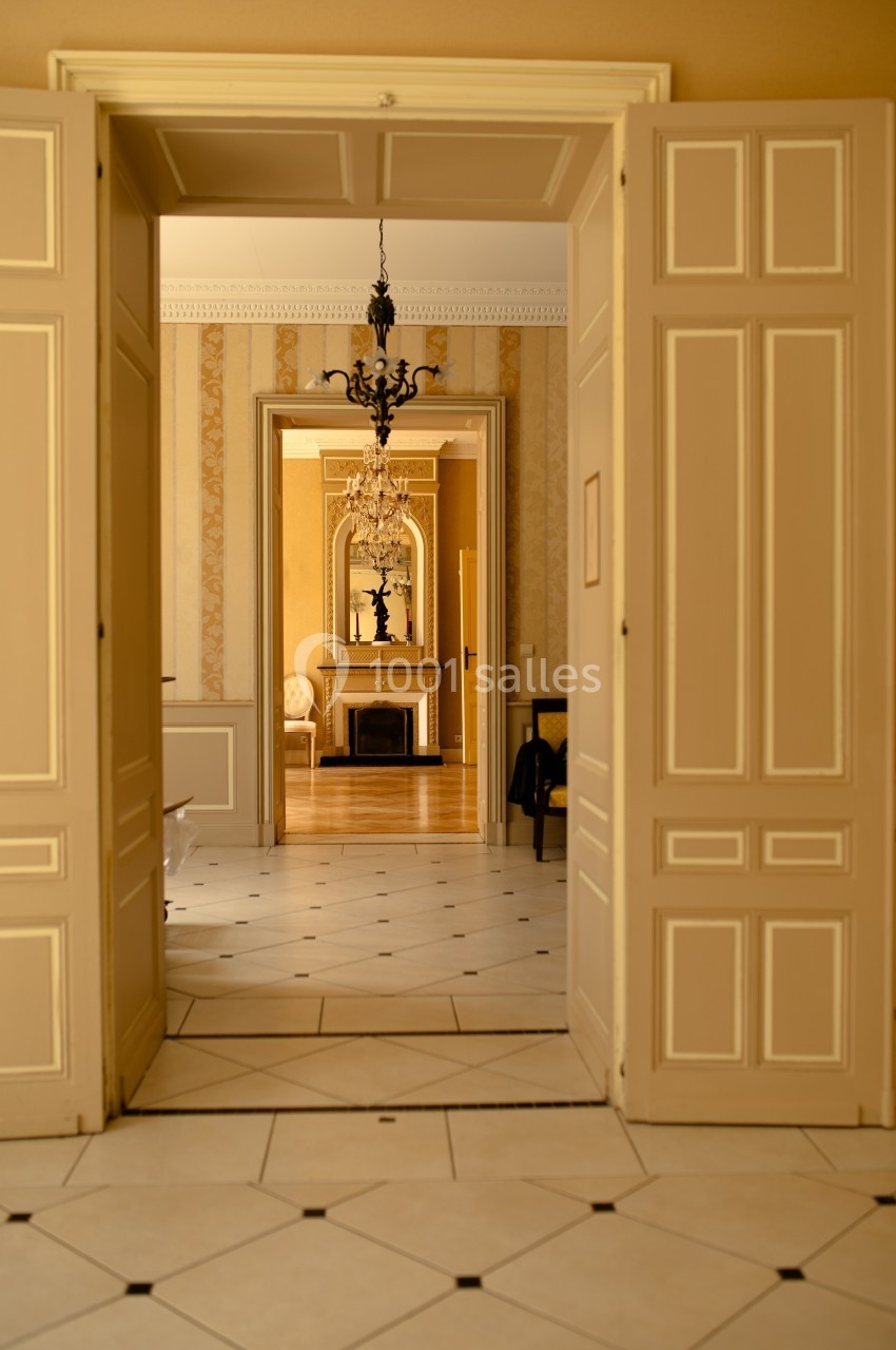 Vue intérieure d'une pièce avec des portes ouvertes, révélant un salon lumineux avec cheminée et lustre.