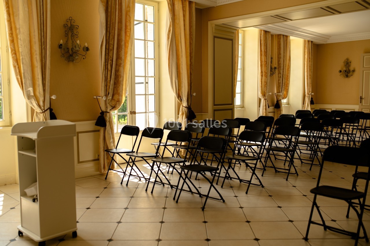 Salle lumineuse avec des chaises pliantes noires alignées devant un pupitre, grandes fenêtres et rideaux dorés.